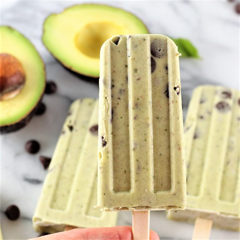 avocado mint chocolate chip creamsicles | Milk & Honey Nutrition Avocado mint chocolate chip popsicle held in front of fresh avocado halves and scattered chocolate chips.