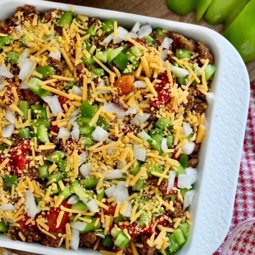 high protein taco dip 4 | Milk & Honey Nutrition White baking dish of layered high protein taco dip topped with diced onions, green peppers, tomatoes and shredded cheddar; tortilla chips nearby.